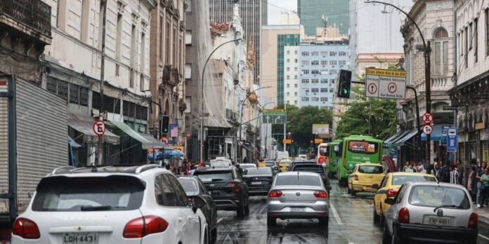 Busy city street after rain with heavy traffic including a white SUV, silver sedan, yellow cabs and a green bus, lined by older buildings and pedestrians on the sidewalks.