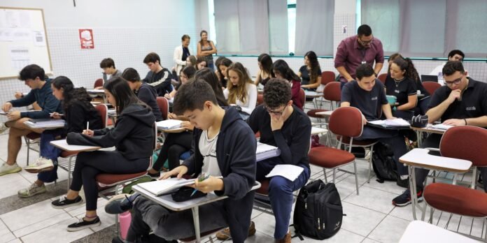 A crowded classroom of students seated at desks, writing in notebooks and studying.