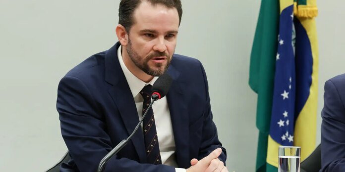 Man in a dark suit and tie speaks into a microphone at a conference, with flags in the background and a glass of water nearby.