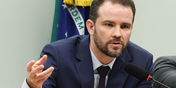 Bearded man in a navy suit speaks into a microphone at a formal press briefing, with a flag in the background.