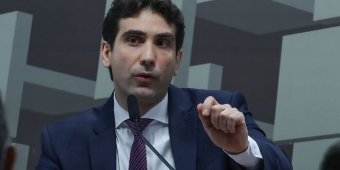 Man in a dark blue suit speaks into a microphone, gesturing with his right hand during a press conference or panel discussion.