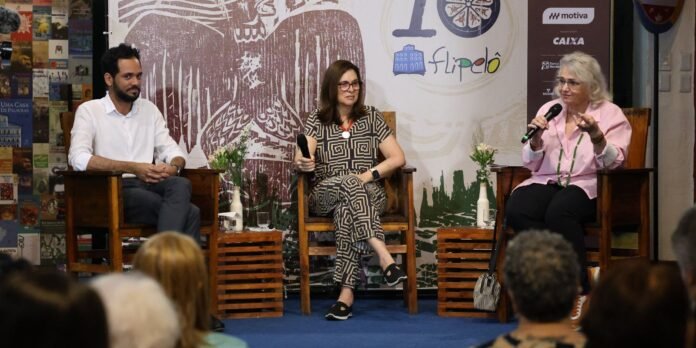 Panel discussion with three speakers on stage: man in white shirt, woman in geometric dress holding a mic, and woman in pink blouse speaking; audience in foreground.