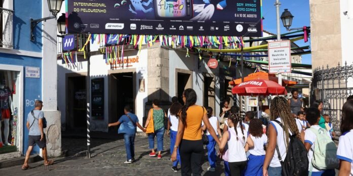 Group of children in white and blue uniforms walk along a cobblestone street under a colorful festival banner with ribbons.