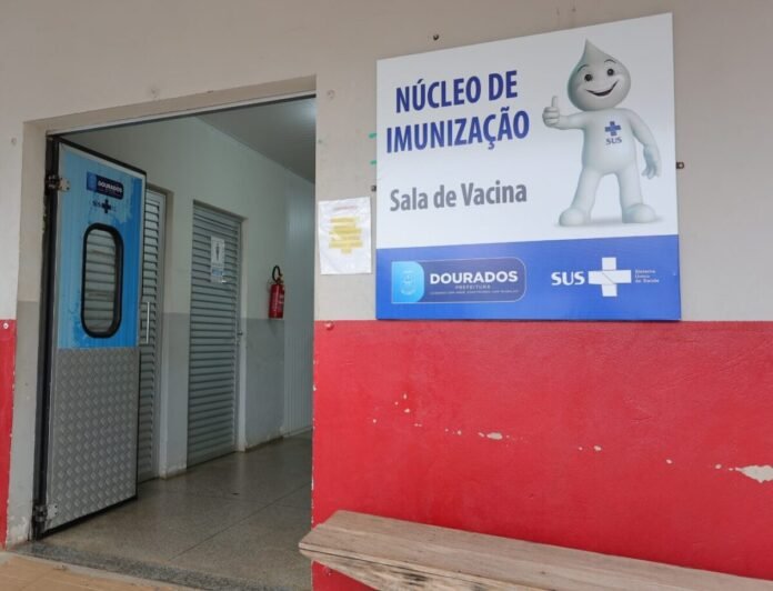 Sign reads Núcleo de Imunização Sala de Vacina with a cartoon water droplet mascot and SUS logo on a clinic wall outside the entrance