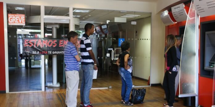 People wait in line inside a bank-like lobby with glass doors; a 'ESTAMOS EM GREVE' banner and a poster reading 'GREVE' are visible.