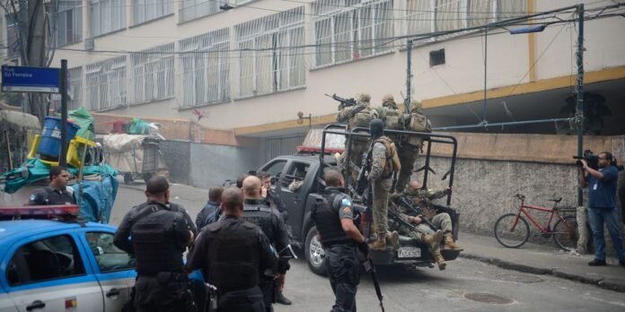 Armed security forces in tactical gear on a pickup truck in an urban street, with a photographer filming nearby.
