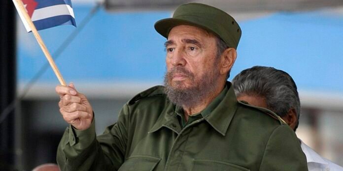 Older man in a green military uniform holds up a small flag on a wooden pole.
