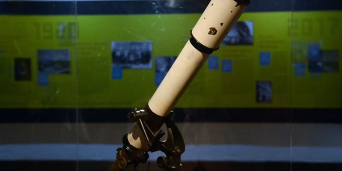 Close-up of a white telescope tube mounted on a metal stand, angled upward against a blurred yellow-green exhibition wall with photos in the background.