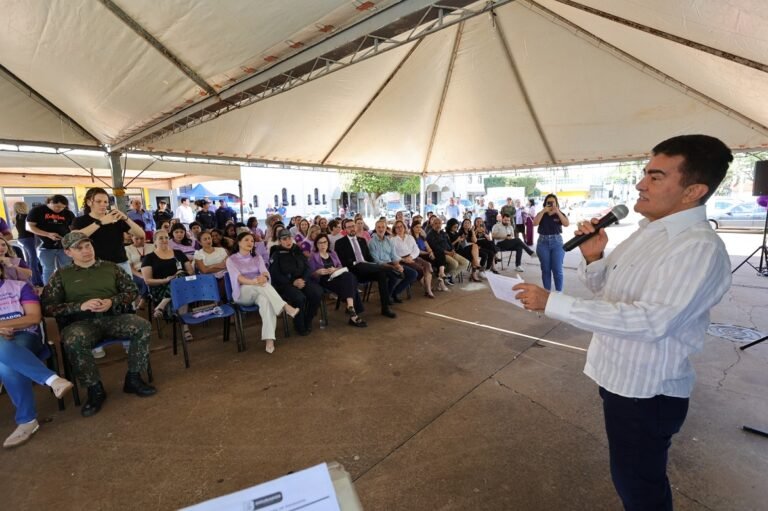 A man in a white striped shirt speaks into a microphone to a seated audience under a large tent at an outdoor event.