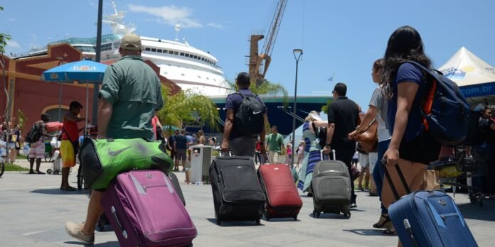 Tourists with rolling suitcases walk toward a large white cruise ship at a sunny port.