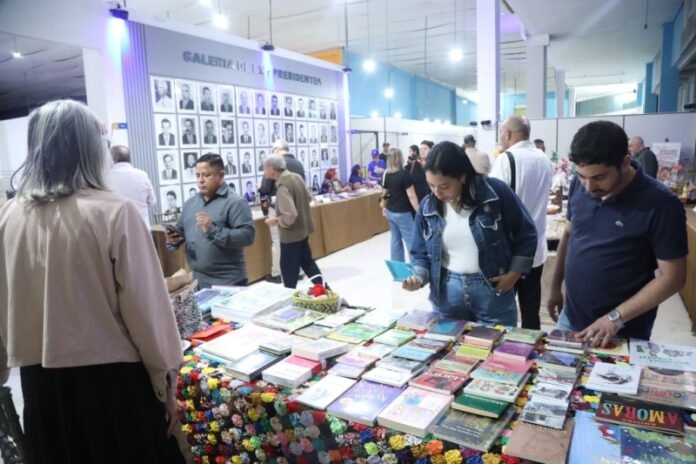People browse a book stall at a lively indoor market, tables stacked with colorful books in the foreground and a portrait wall in the background.