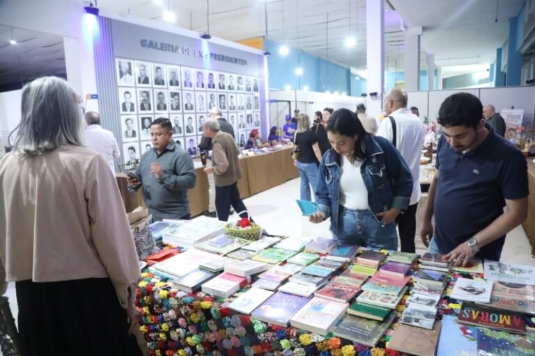 People browse a book stall at a lively indoor market, tables stacked with colorful books in the foreground and a portrait wall in the background.