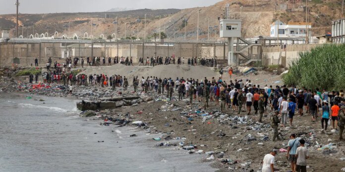 Long line of people walking along a polluted rocky beach beside a high-security compound with barbed wire and a watchtower in the background, guarded by fences and security towers.