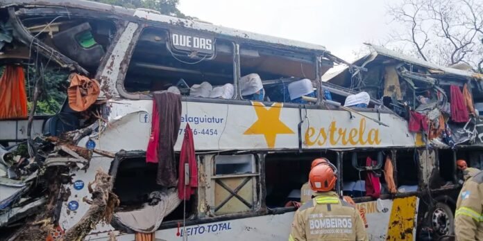 Damaged bus wreck with exposed interior, torn panels and hanging clothes; firefighters in orange gear stand nearby.
