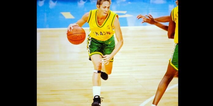 Female basketball player in yellow and green Brazil uniform dribbles the ball while a defender reaches in on a wooden court.