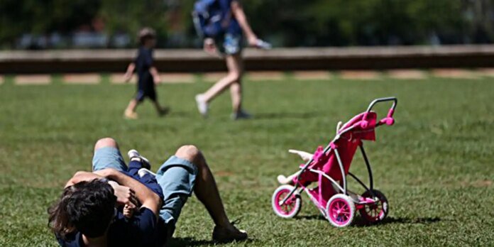 Man lying on grassy park field with a pink toy stroller nearby as kids play in the background.