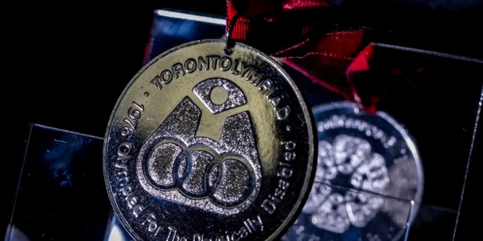 Close-up of a gold medal hanging from a red ribbon against a dark background, featuring interlocking rings emblem and 'Toronto' text.