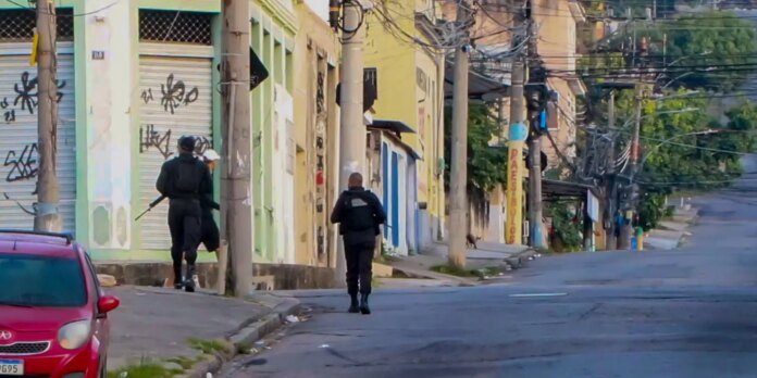 Two armed officers in black gear walk down a graffiti-covered urban street from behind. An orange car is parked on the left.