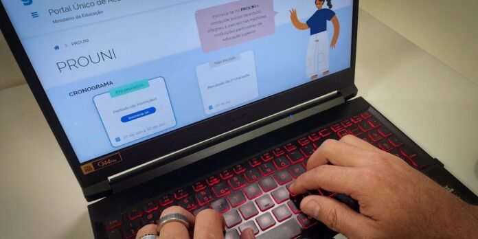 Close-up of a person typing on a black laptop with red backlit keyboard; screen shows a Portuguese education portal (PROUNI) with a schedule card.