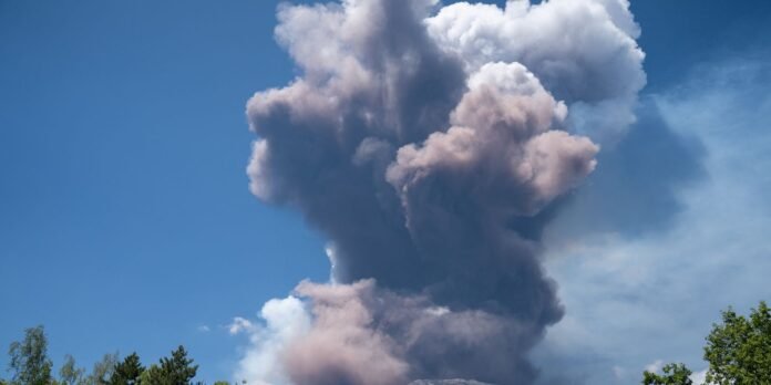 Tall ash and smoke plume rising into a clear blue sky, with treetops at the bottom suggesting an eruption in the distance.