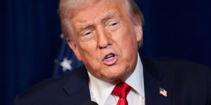 Close-up of a man with light hair in a dark suit and red tie speaking into a microphone, wearing a small American flag pin on his lapel.