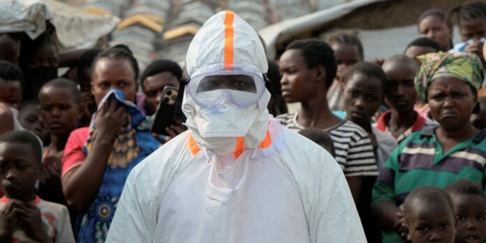 Health worker in full white protective suit and goggles stands in front of a crowd of onlookers outdoors, with bright orange accents on the suit neck and hood.