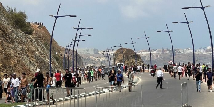 Crowded coastal promenade with pedestrians walking along a curved road, blue streetlamps, and rocky cliff on the left with a distant cityscape across the bay.