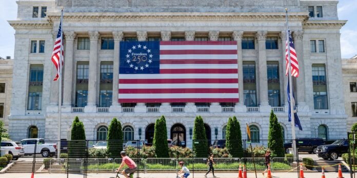 Front of a marble government building with a large American flag banner reading 'Freedom 250' between tall columns; pedestrians and cars in front.