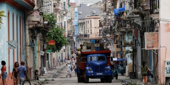 Narrow street in a colorful city, a blue vintage truck loaded with crates in the middle, with pedestrians and pastel buildings on both sides.