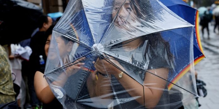 Two people share a large, transparent umbrella with a rainbow-trim edge on a rainy city street, raindrops glistening on the plastic surface (scene in the background).