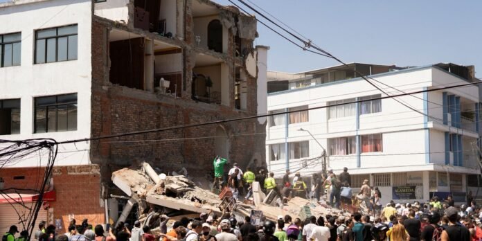 Crowd of rescue workers and onlookers atop a collapsed brick building with debris in the street.