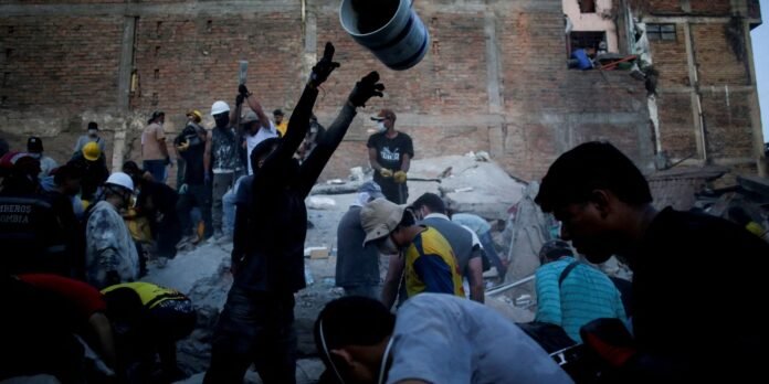 Crowd of rescue workers in helmets and masks sift through debris at a collapsed building by a brick wall.