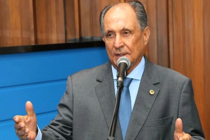 Older man in a gray suit and blue tie speaks into a microphone, gesturing with his hands in a wood-paneled room.