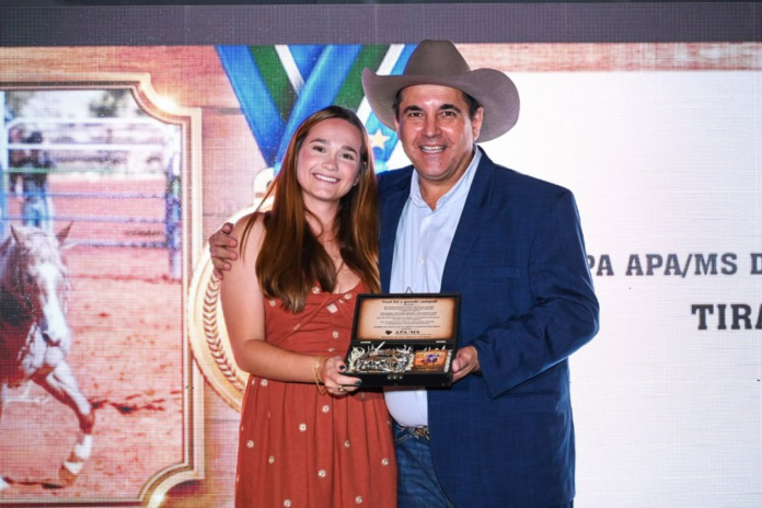 Two people pose on stage holding an award plaque, smiling at the camera. The man wears a blue blazer and a cowboy hat.
