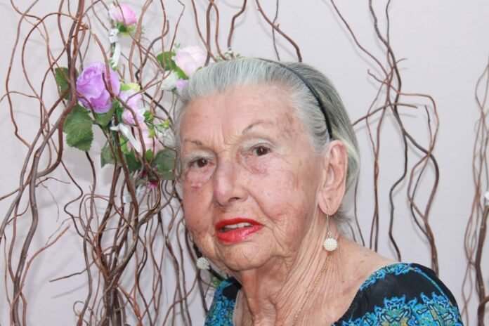 Portrait of an elderly woman with silver hair, wearing a blue patterned top, red lipstick, and pearl earrings, posed beside decorative branches and flowers.