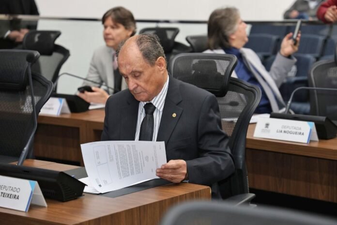 Man in a dark suit reads papers at a wood desk in a formal meeting room in progress of a session as others sit behind him.