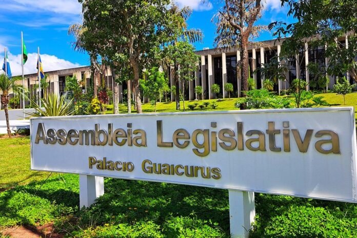 Large white sign with bronze letters reading Assembleia Legislativa and Palacio Guaicurus, set in a green lawn with trees and blue sky behind it