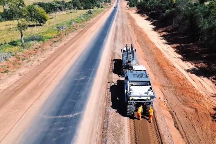 Aerial view of a heavy road construction truck laying new asphalt on a rural highway, with workers nearby.