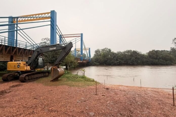 Yellow Deere excavator parked on a rocky riverbank beside a blue steel bridge over a calm river.