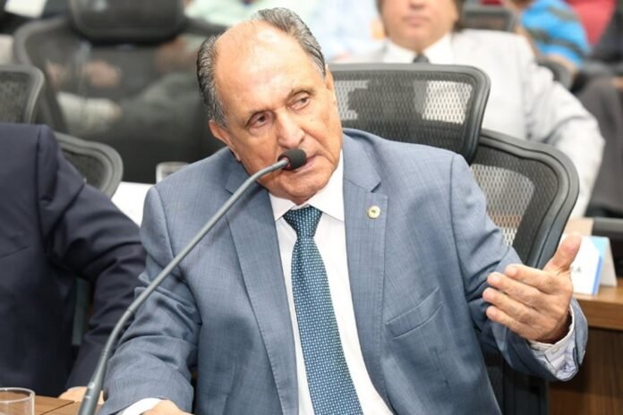 Older man in a blue suit and patterned tie speaks into a microphone at a formal meeting, gesturing with his right hand.