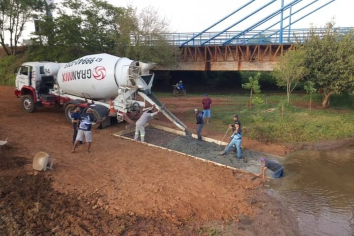 Construction crew pours wet concrete from a mixer truck into a trench along a riverbank near a blue bridge.