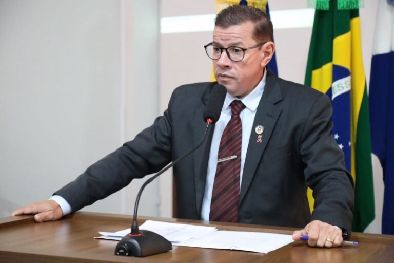 Man in a dark suit and glasses speaks into a microphone at a desk during a formal briefing, with papers in front and flags behind him.
