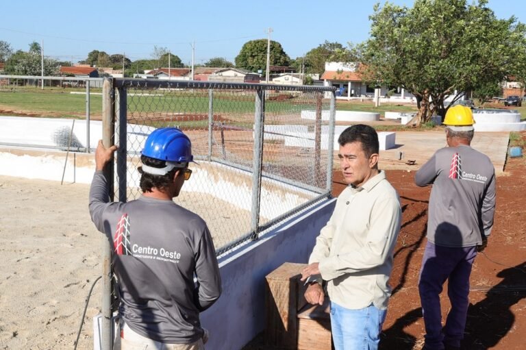 Three workers at a construction site by a chain-link fence, one in a blue helmet talking to a man in a light shirt and another worker nearby.