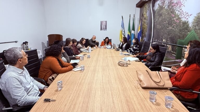 Diverse group seated around a long conference table in a meeting room, with flags and a portrait on the wall behind them.