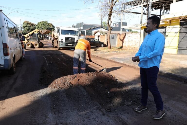 Worker in orange shirt shovels dirt on a road under construction; a man in a blue shirt watches nearby.