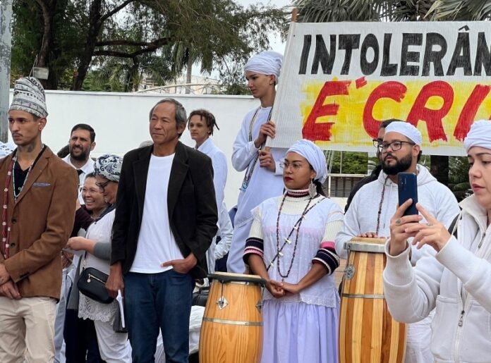 Group of people in white traditional clothing with drums, standing behind a large protest sign; a man in a blazer stands front center, others watch or film, some wear headwraps.