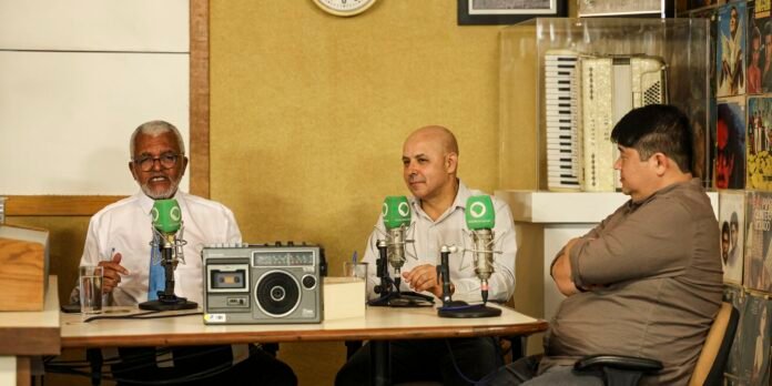 Three men sit at a table in a radio studio, speaking into green microphone flags during a live podcast.