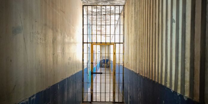 Narrow prison hallway seen through a tall metal gate, with barred doorway at the end and blue lower walls.