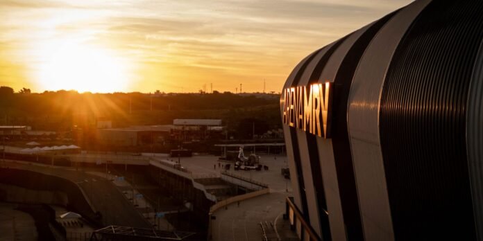 A curved modern building with a glowing ARENA MRV sign at sunset, casting warm light over the plaza below.