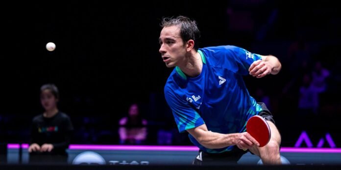 Male table tennis player in a blue shirt lunges to strike a white ball with a red paddle in a dim arena, mid-match action.